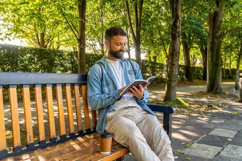 Man with a beard sitting on a wooden park bench, focused on reading a book with a coffee cup beside