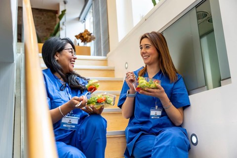 Two smiling female healthcare professionals in scrubs sitting on hospital stairs, eating fresh