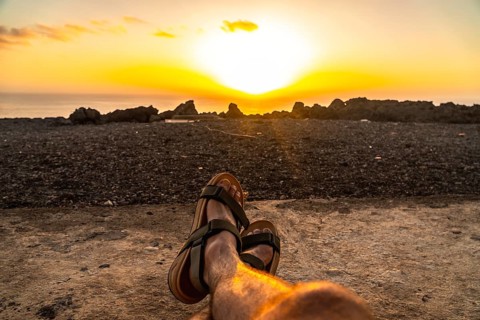 Person's feet wearing sandals relaxing on the beach sand, watching the golden orange sunset over