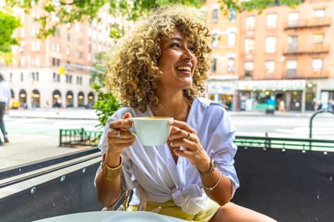 Woman with curly hair smiling and laughing, holding a coffee cup, sitting at an outdoor cafe on a