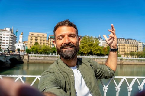 Bearded man smiling and taking a selfie while standing on a bridge, enjoying his travel and