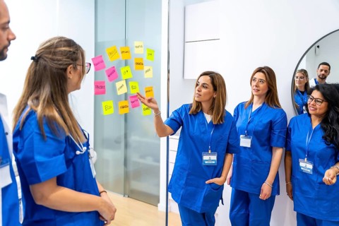 Medical team in blue scrubs gathered at a glass board with colorful sticky notes, collaboratively