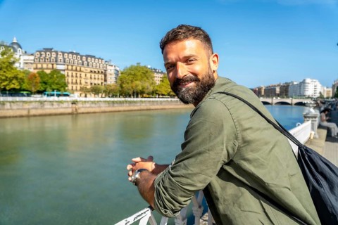 Young man with a beard smiling and looking at camera, leaning on a bridge railing with a city river