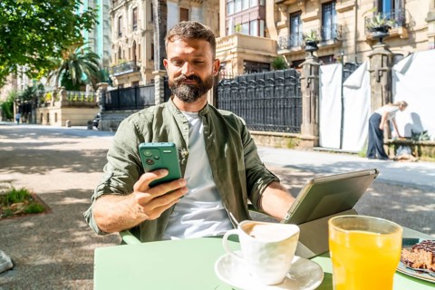Bearded man working remotely with his smartphone and tablet while enjoying breakfast and coffee at