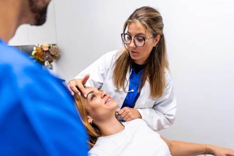 Young doctor in white coat and glasses examines a female patient's forehead, assessing skin