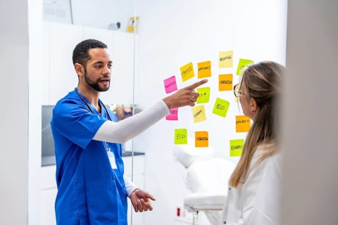 Healthcare team in scrubs and white coats brainstorm and prioritize ideas on a glass board with