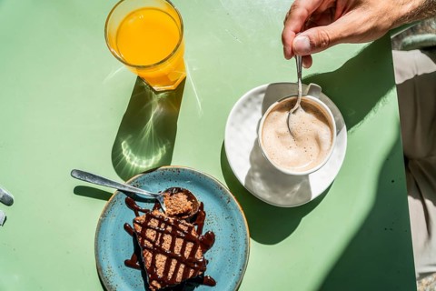 Person enjoying a sunny outdoor breakfast, stirring a cup of frothy coffee next to a slice of