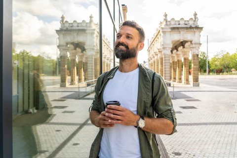Smiling man enjoying a hot drink in a reusable cup while walking through a modern urban environment