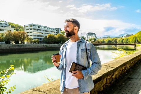 Young man holding a disposable coffee cup and a book, enjoying a relaxed morning walk along a city