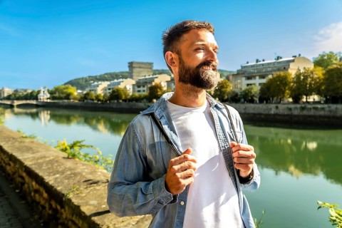 Smiling bearded man with a backpack strolls along a sunny riverside promenade in the city, enjoying