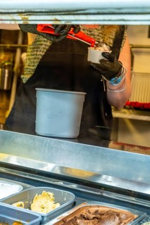 Worker wearing black gloves and an apron scooping a portion of ice cream from a display freezer