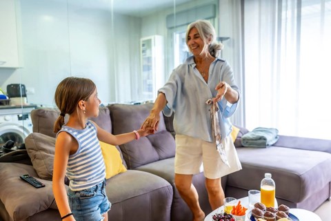 Grandmother holding granddaughter's hand, looking joyful while dancing in a bright living room at