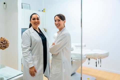 Two smiling female doctors or beauticians wearing white coats, posing confidently in a contemporary