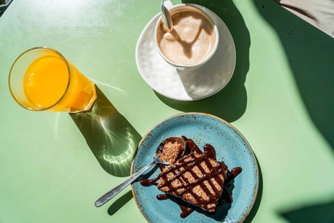 Breakfast spread on a green outdoor table with coffee, orange juice and cake, bright harsh sunlight