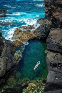 Woman floating on her back in a serene turquoise natural pool surrounded by dark volcanic rocks,