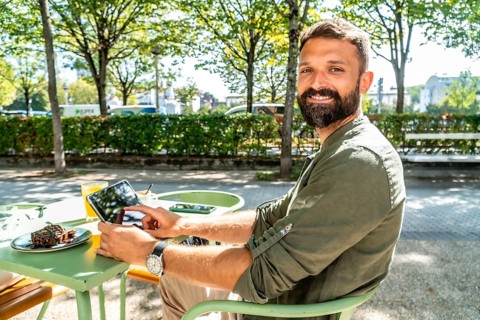 Man with a beard smiling at the camera, using a digital tablet at a green outdoor cafe table with a
