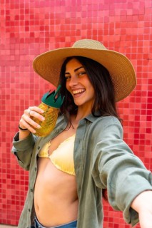 Young woman broadly smiling, wearing a straw hat and bikini top under an open shirt, holding a