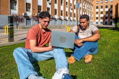 Two diverse young men sitting on a university lawn, collaborating on a laptop, smiling and studying
