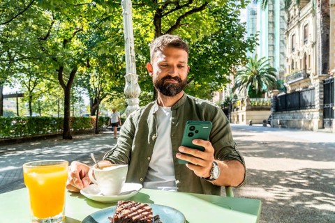 Bearded young man in a casual shirt enjoys breakfast at an outdoor cafe, checking his smartphone