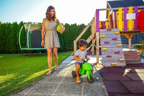 Mother holding ball walking and watching her son riding toy motorbike in backyard on summer