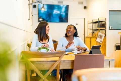 Two young female friends enjoying a coffee break, laughing and talking together at a table in a