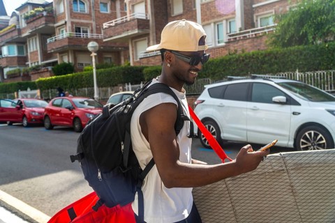 Young man wearing sunglasses, a cap, and a backpack, walking on a city street while engaging with