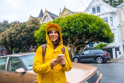 Woman in yellow raincoat stands on a san francisco street, looking up with smartphone in hand amid