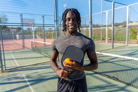 Young athletic black man standing on an outdoor pickleball court, holding a paddle and ball,