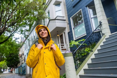 Young woman wearing a yellow raincoat and backpack, looking up while walking along a street lined