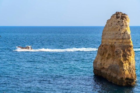 Motorboat full of tourists sailing near a majestic sea stack in the beautiful algarve region of