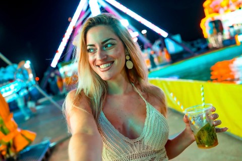 Blonde smiling woman taking selfie while drinking cocktail at fair at night