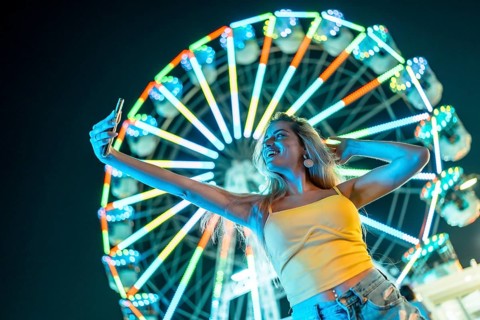 Happy woman taking selfie next to illuminated ferris wheel at night in an amusement park