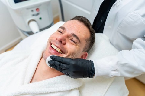 Man smiling while a professional applies a facial skincare product to his face with a cotton pad,