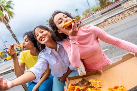 Chinese woman taking selfie while eating pizza with friends sitting in the street in the city