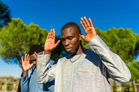 Two young senegalese men wearing traditional clothing are praying outdoors with their hands raised,