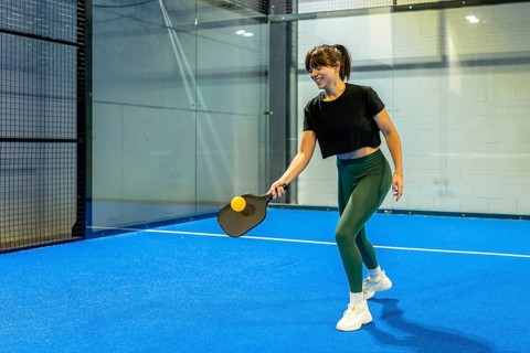 Smiling woman playing pickleball, hitting a yellow ball with a black paddle on a blue indoor court,