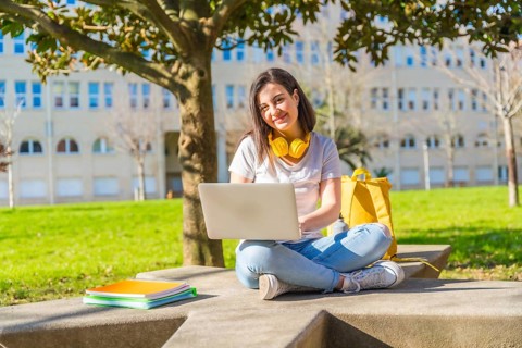 Portrait of a cute smiling woman using laptop to study outside the university while wearing