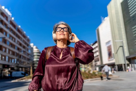 Grey haired woman with eyeglasses and headphones standing outdoors, enjoying music with eyes closed