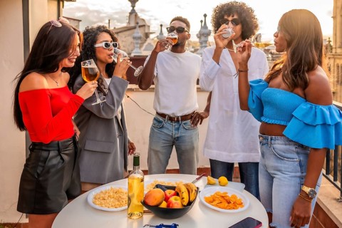 Group of diverse friends drinking wine and eating snacks in rooftop during sunset party
