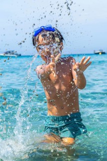 Joyful boy wearing blue swimming goggles and green shorts, playing and splashing water in the clear