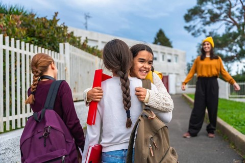 Young girls wearing backpacks happily embracing and laughing at school while an adult woman watches