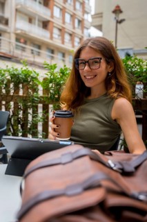 Young woman wearing glasses smiling while enjoying a hot coffee outdoors at a city cafe with a