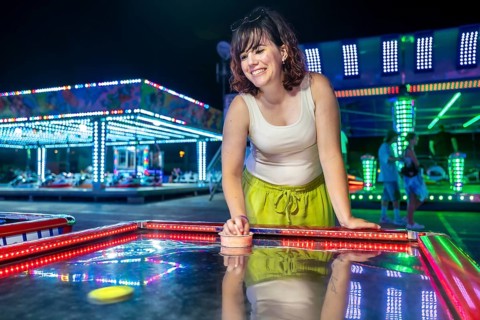 Happy young woman laughing while playing air hockey at a vibrant night amusement park, enjoying fun