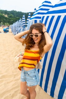 Young woman standing on a sandy beach, laughing with hands in her hair, wearing a colorful striped