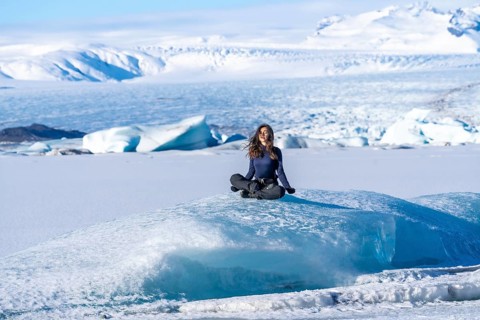 A woman is sitting on a large block of ice. The ice is surrounded by snow and the woman is looking