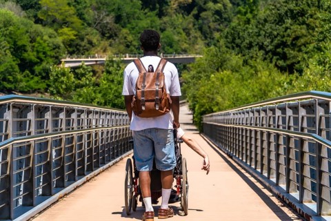 Person pushes another individual in a wheelchair across a modern bridge surrounded by lush green