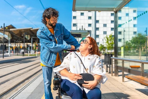 Young man joyfully pushing a smiling woman in a wheelchair at a modern tram station, embodying