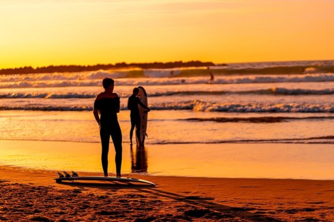 A man stands on the beach with his surfboard, watching the waves. The sky is orange and the water