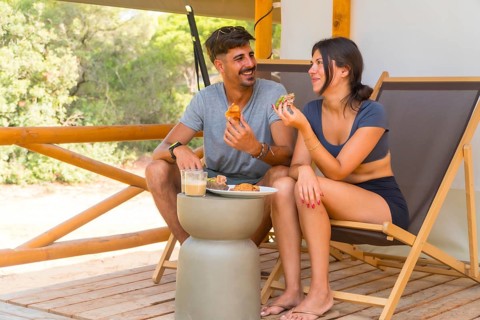 Young couple smiling and having breakfast on the wooden terrace of a glamping tent, surrounded by