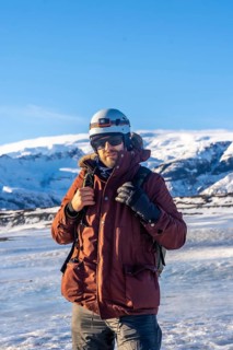 A man wearing a red jacket and a helmet stands in the snow. He is holding a camera and has a